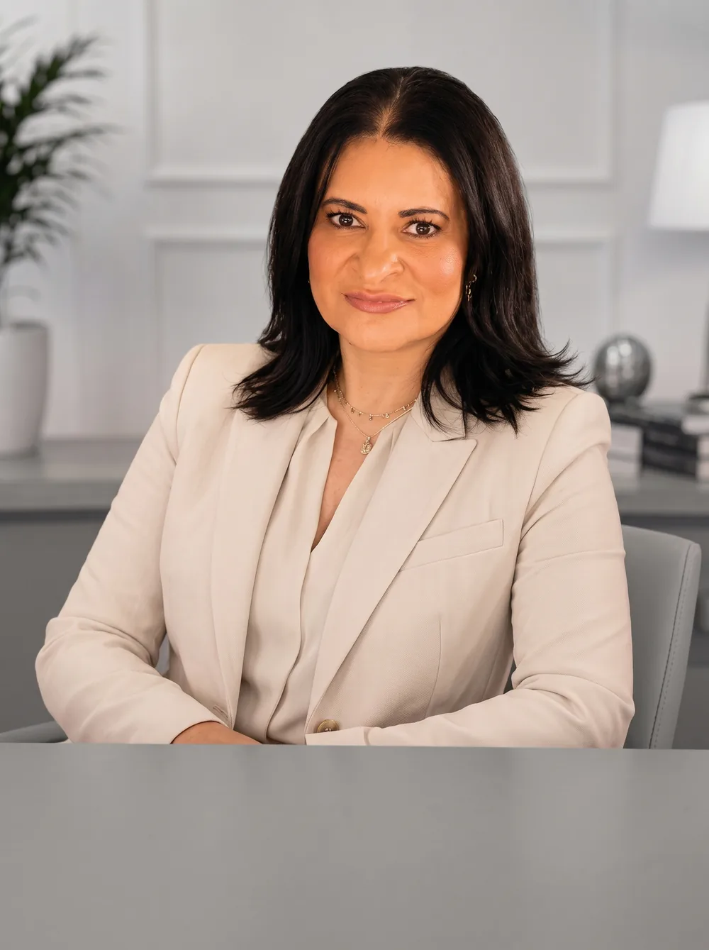 Dra. Willia Ferreira, advogada, de blazer claro e cabelo escuro na altura dos ombros, sorrindo sentada à mesa do escritório.